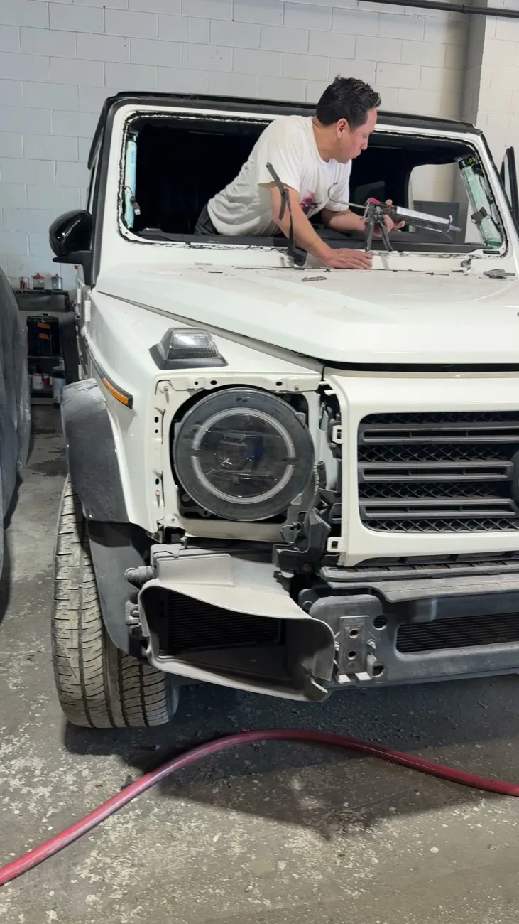 Exclusive Collision & Repair technician running urethane through the windshield opening of a white Mercedes G-Class with the headlight surround and bumper stripped off in the Cypress Hills shop in Brooklyn