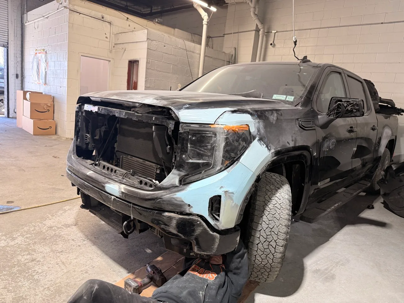 Technician working underneath a black GMC Sierra crew cab with the front bumper off and a primered fender, exposing the core support during body-on-frame structural repair at Exclusive Collision & Repair in Brooklyn, NY
