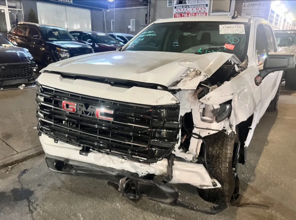 White GMC Sierra pickup with the front bumper, grille and headlight destroyed and the intake number marked on the windshield, parked outside Exclusive Collision & Repair on Marginal Street in Brooklyn after a tow